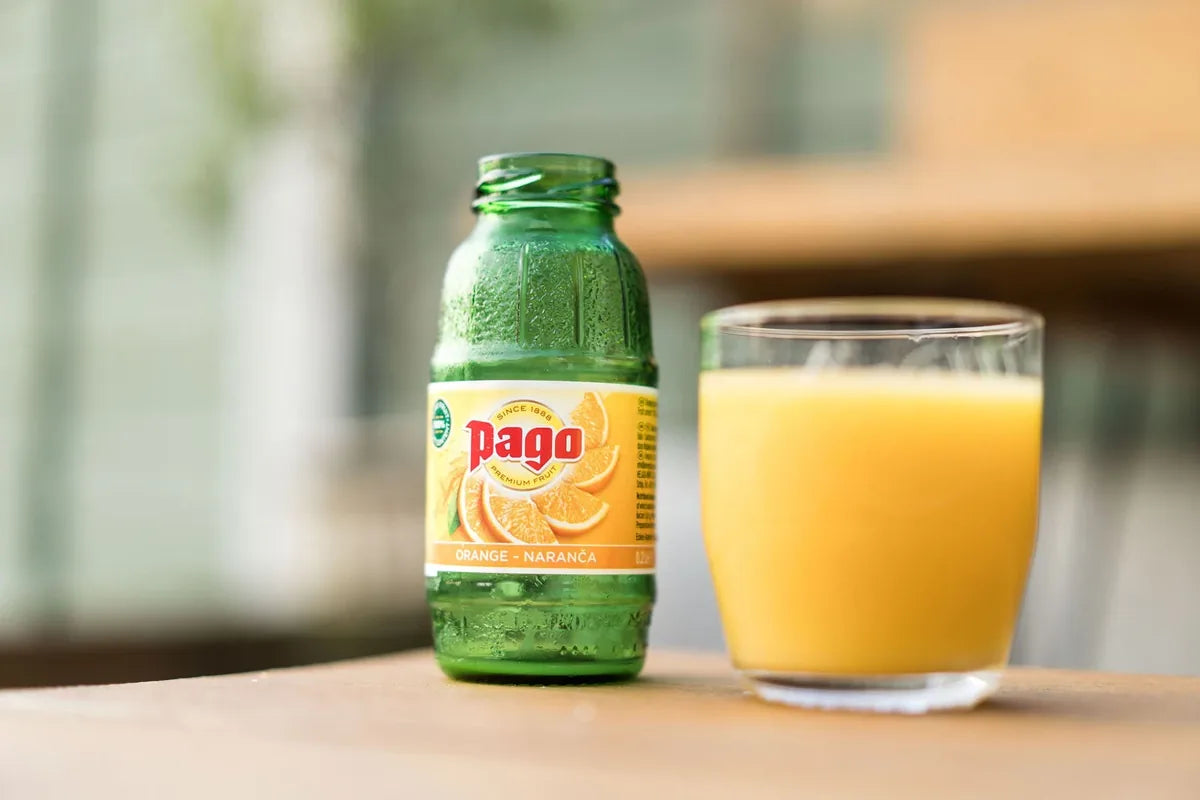 Een geopend groen flesje Pago Orange naast een glas sinaasappelsap